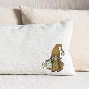 May include: A white throw pillow with a brown and white embroidered gnome holding a steaming cup of coffee. The gnome is wearing a brown hat and a brown coat.