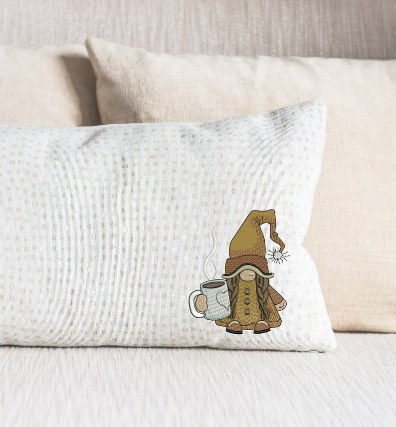 May include: A white throw pillow with a brown and white embroidered gnome holding a steaming cup of coffee. The gnome is wearing a brown hat and a brown coat.