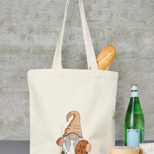 May include: A white canvas tote bag with a brown and gray gnome holding a button and a needle. The gnome is embroidered on the bag.