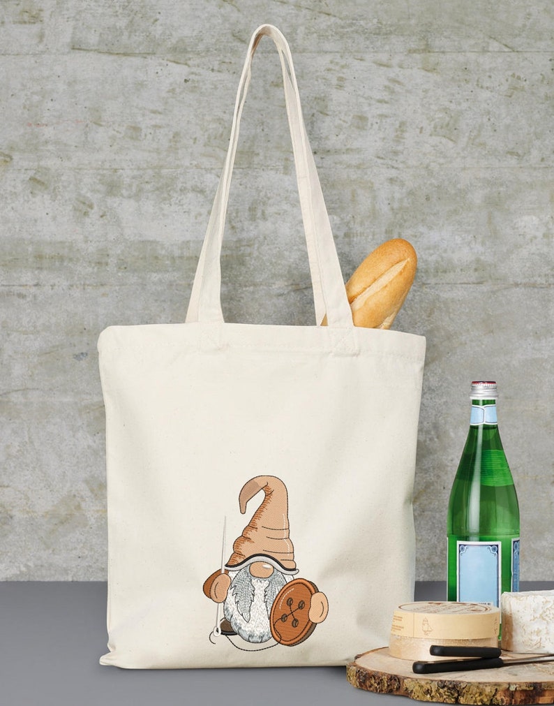 May include: A white canvas tote bag with a brown and gray gnome holding a button and a needle. The gnome is embroidered on the bag.