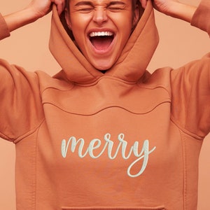 May include: A woman wearing a terracotta orange hoodie with a white embroidered "merry" on the front. She is pulling the hood up over her head and laughing.