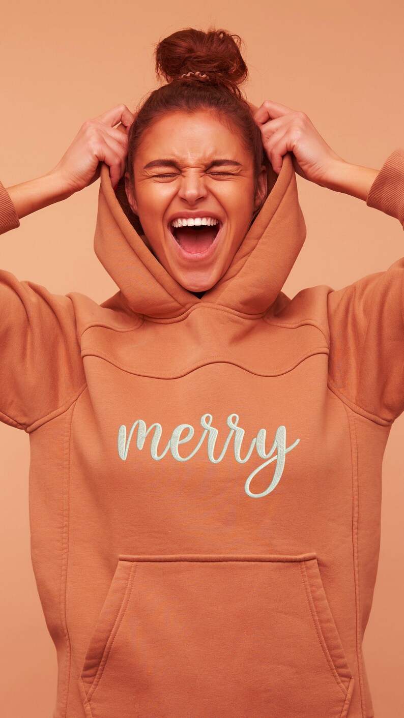 May include: A woman wearing a terracotta orange hoodie with a white embroidered "merry" on the front. She is pulling the hood up over her head and laughing.