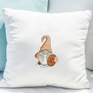 May include: A white pillow with a brown and white gnome holding a brown button. The gnome has a brown hat and a white beard.