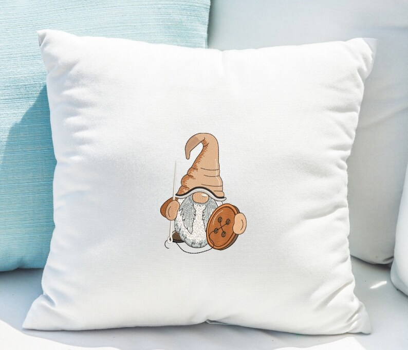 May include: A white pillow with a brown and white gnome holding a brown button. The gnome has a brown hat and a white beard.