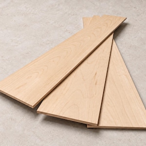 May include: Three rectangular wooden boards arranged in a fan-like overlapping pattern. Each board features a light tan color with visible natural wood grain patterns running lengthwise across the flat, smooth surfaces of the planks.