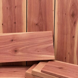 May include: A collection of rectangular cedar wood planks featuring a reddish-brown hue with visible natural wood grain patterns and dark knots. The boards are arranged in a stacked formation with some standing vertically and others lying horizontally.