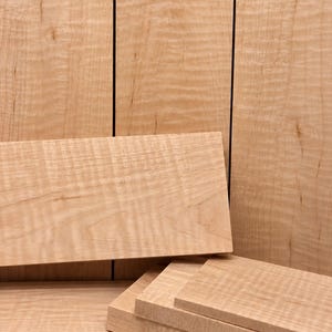 May include: A collection of light-colored, rectangular wooden boards featuring a distinct wavy, rippled grain pattern. The boards are arranged in a stack with several pieces lying horizontally and others standing vertically in the background.