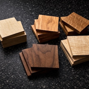 May include: A collection of square wooden coasters in various wood tones, including light, medium, and dark shades. The coasters are stacked in small piles, showcasing the natural grain and texture of the wood. The coasters are arranged on a dark, speckled surface.