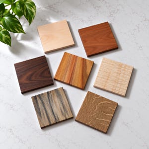 May include: Seven square wooden coasters arranged in a cluster, each displaying a distinct wood grain pattern and color, ranging from light cream and pale maple to rich reddish-brown, dark walnut, and textured grey-brown wood tones.