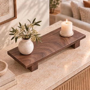 May include: A rectangular dark brown wooden riser with four short block feet. On the surface sits a small, round, speckled off-white ceramic vase containing green leafy stems and a white pillar candle resting on a circular stone coaster.
