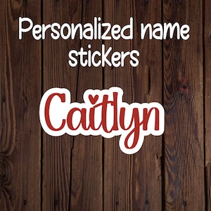May include: A personalized name sticker with the name "Caitlyn" in a red, cursive font, outlined in white. The sticker is on a wooden background. The text "Personalized name stickers" is above the name in white.