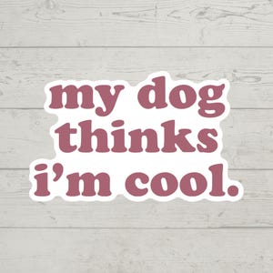 Puede incluir: Una pegatina malva con la frase "my dog thinks i'm cool" en una fuente redondeada con contorno blanco. El fondo es de madera clara.