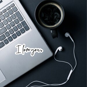 May include: A laptop computer with a white sticker that says "I love you."  The laptop is on a black surface with a cup of coffee and white earbuds.
