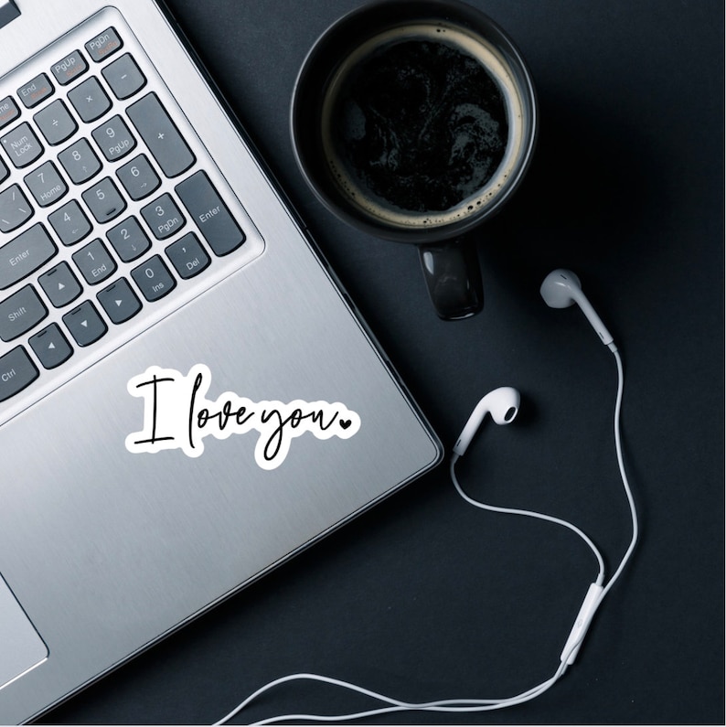 May include: A laptop computer with a white sticker that says "I love you."  The laptop is on a black surface with a cup of coffee and white earbuds.
