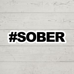 May include: Black and white sticker with the text "#SOBER" on a white wood background.