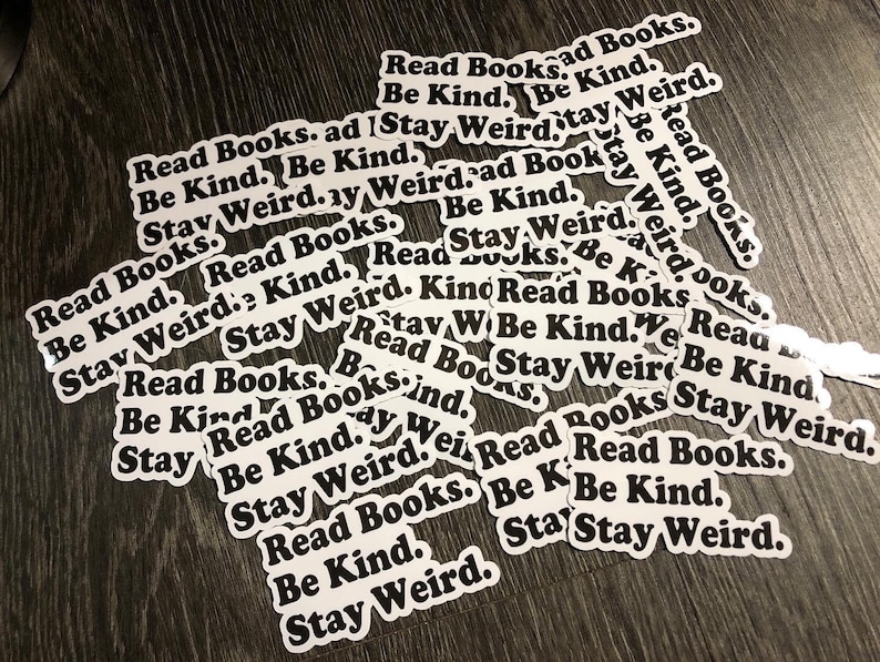 May include: A pile of white stickers with black text that reads "Read Books. Be Kind. Stay Weird." The stickers are scattered on a wooden surface.