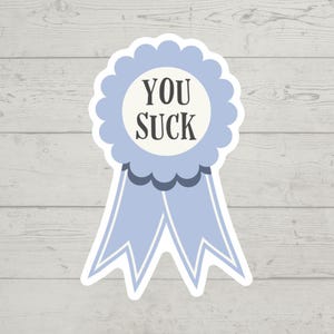 Puede incluir: Una pegatina de cinta azul claro con las palabras "YOU SUCK" en texto negro. La pegatina tiene un borde festoneado y dos colas de cinta. El fondo es un patrón de vetas de madera clara.