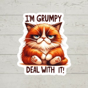 Peut inclure: Un chat tabby orange à l'air grognon avec des yeux bleus est assis les pattes croisées et fait la moue. Le texte "I'm Grumpy Deal With It!" est écrit au-dessus et en dessous du chat.