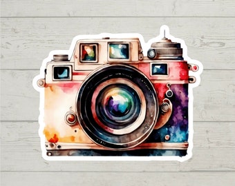 Può includere: Un'illustrazione ad acquerello di una macchina fotografica vintage con uno schema di colori rosso, bianco e blu. La macchina fotografica ha un obiettivo grande e una varietà di quadranti e pulsanti.