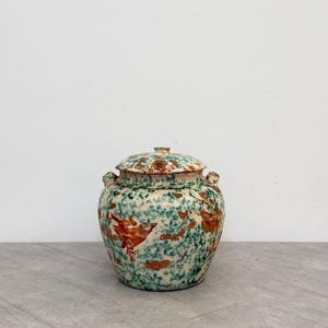 May include: A ceramic jar with a lid featuring a mottled pattern of teal and green speckles layered with patches of burnt orange and brown. The jar has a rounded body, a short neck, and two small handles on the sides, showcasing a textured finish.