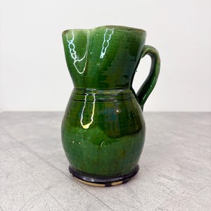 Puede incluir: Jarra de cerámica verde hecha a mano, con base bulbosa y borde festoneado. La jarra presenta un asa curva y un acabado brillante, mostrando variaciones en el esmalte verde. La base es marrón oscuro. Una pieza decorativa para la cocina.