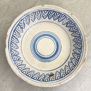 Op de afbeelding: Een wit keramiek bord met een blauw-wit ontwerp. Het bord heeft een gekrakelde glazuur en een blauwe ring rond het midden. De rand van het bord is versierd met een herhalend patroon van blauwe driehoeken.
