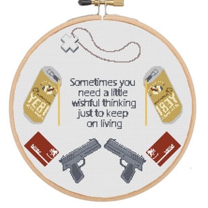 Peut inclure: Cerceau de broderie au point de croix avec un fond blanc et une variété d'images colorées, dont deux canettes de bière, deux armes à feu, deux cartes rouges et un collier. Le texte "Sometimes you need a little wishful thinking just to keep on living" est brodé en fil noir.