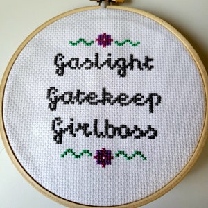 Peut inclure: Cerceau de broderie blanc avec inscription brodée en noir "Gaslight Gatekeep Girlboss" avec des accents floraux verts et violets.