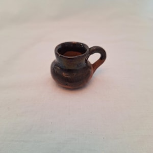 May include: A small, handcrafted ceramic mug with a dark brown exterior and a lighter brown interior. The mug features a rounded body, a small handle, and a slightly flared rim. The mug is approximately 2 inches tall.