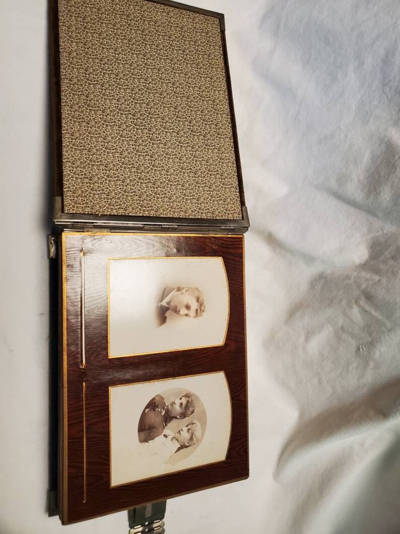 May include: A vintage photo album with a brown leather cover and gold accents. The album is open to reveal two black and white photos of people.