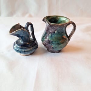 May include: Two small ceramic pitchers. The pitcher on the left is blue with a unique handle and spout. The pitcher on the right is a rounded shape with a handle and is glazed in shades of green, purple, and brown.