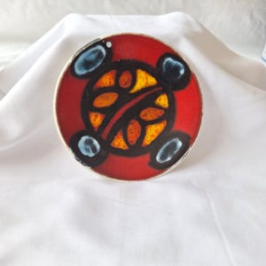 May include: A round, red ceramic dish with a black, orange, and yellow abstract design. The dish has a white rim and four blue and black accents. The design is centered on the dish.
