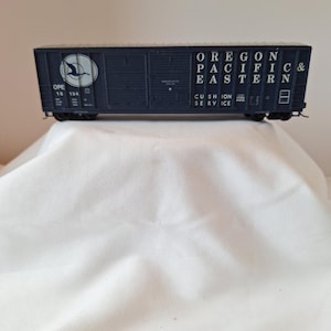 May include: A dark blue model railroad boxcar featuring the text OREGON PACIFIC & EASTERN and CUSHION SERVICE. The side displays a circular logo with a bird silhouette, the number OPE 15 134, and various technical markings in white lettering.
