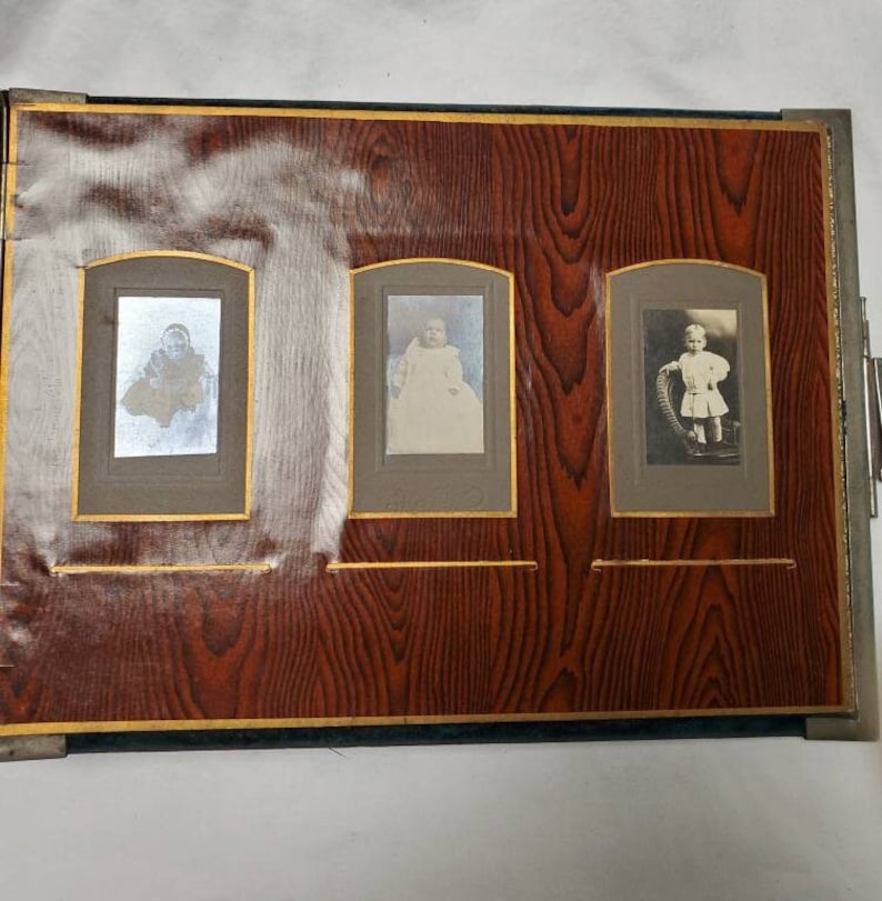 May include: A vintage photo album with a dark brown wood-grain cover and three photo slots. Each slot has a gold trim and contains a black and white photo of a baby.