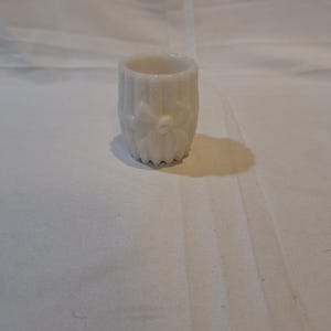 May include: A small, white, milk glass cup with a ribbed design and a raised bow at the top.