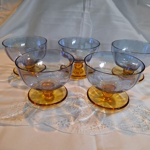 May include: Set of six clear glass dessert cups with a blue iridescent tint and amber colored bases. The cups are arranged on a white lace tablecloth.
