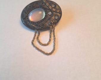 May include: An oval-shaped brooch with a silver-toned metal frame. The brooch features a central, iridescent, oval-shaped stone and is accented with small clear stones and a dangling gold-colored chain. The design has an antique or vintage aesthetic.