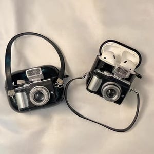 Puede incluir: Dos fundas protectoras negras para auriculares inalámbricos, cada una diseñada como una cámara vintage con lente plateada, flash y correa negra. Una funda está cerrada, mientras que la otra está abierta, mostrando auriculares blancos dentro.