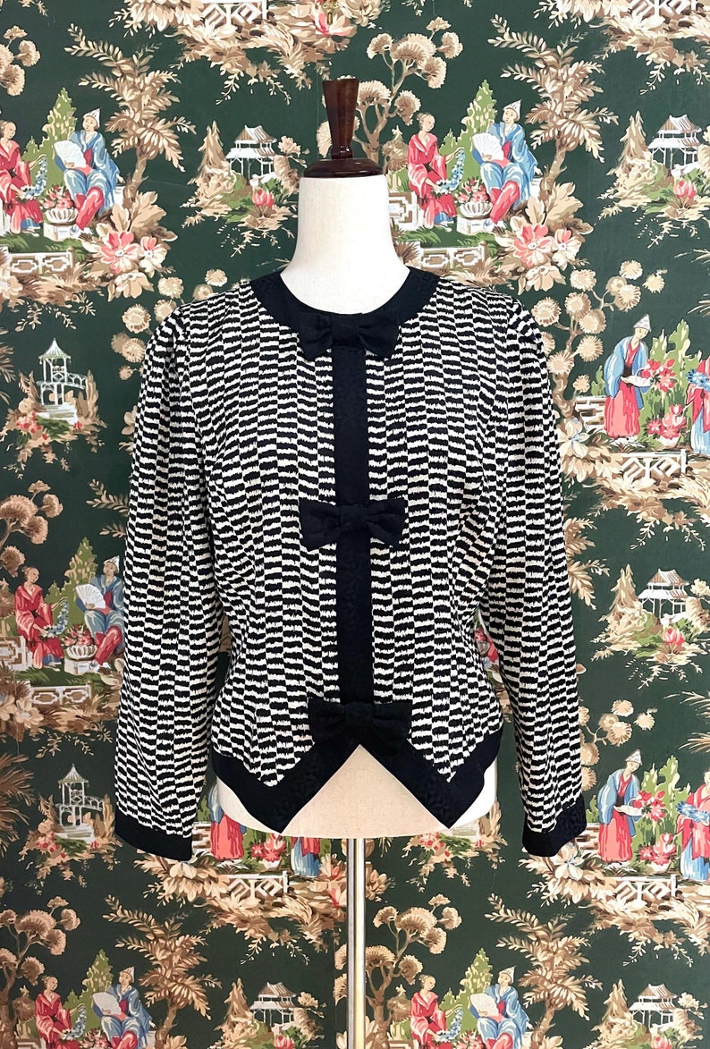 May include: A black and white striped blouse with a black bow at the neckline. The blouse has long sleeves and a fitted silhouette.