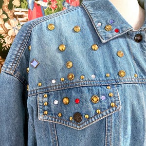 Può includere: Giacca di jeans azzurra decorata con borchie dorate, dettagli a spirale e strass colorati. La giacca presenta un colletto classico, chiusura con bottoni e una tasca sul petto. Un capo unico ed elegante.