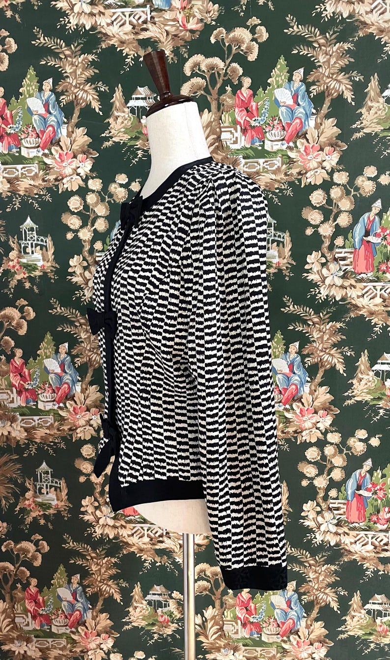 May include: A black and white striped cardigan with a black bow detail. The cardigan is on a white mannequin against a green and white floral patterned background.
