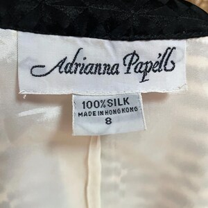 May include: White tag with black text that reads "Adrianna Papell" and "100% Silk Made in Hong Kong 8". The tag is attached to a white garment with a black trim.