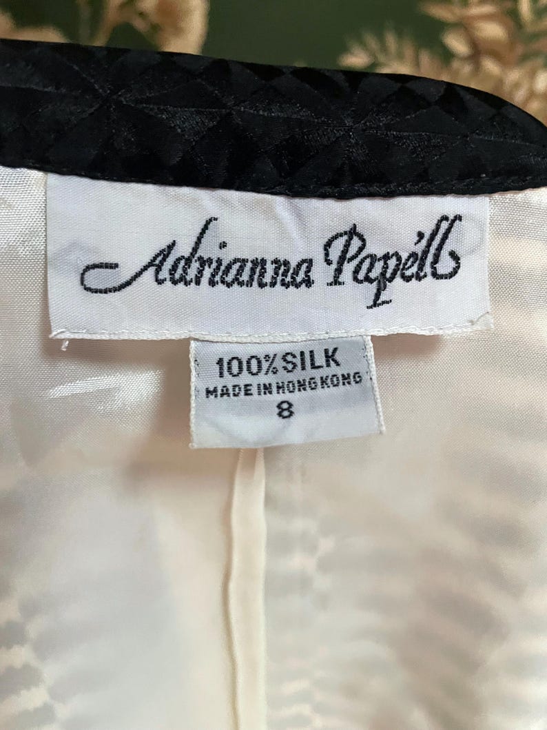 May include: White tag with black text that reads "Adrianna Papell" and "100% Silk Made in Hong Kong 8". The tag is attached to a white garment with a black trim.