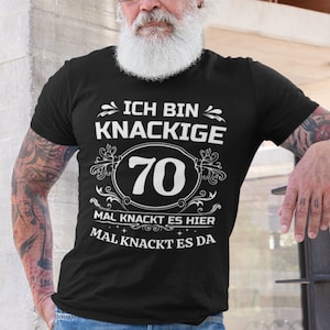 Op de afbeelding: Een zwart T-shirt met witte tekst die "Ich bin knackige 70 Mal knackt es hier Mal knackt es da" zegt.