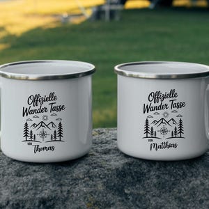 Puede incluir: Dos tazas de camping de esmalte blanco con texto negro y un dise&ntilde;o de monta&ntilde;a y br&uacute;jula. Las tazas tienen un borde y un asa plateados. Una taza dice "Offizielle Wander Tasse von Thomas" y la otra "Offizielle Wander Tasse von Matthias."