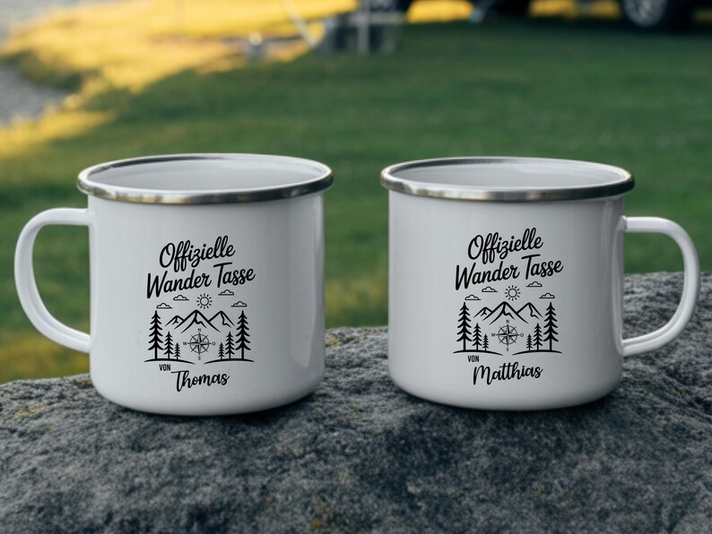 Puede incluir: Dos tazas de camping de esmalte blanco con texto negro y un dise&ntilde;o de monta&ntilde;a y br&uacute;jula. Las tazas tienen un borde y un asa plateados. Una taza dice "Offizielle Wander Tasse von Thomas" y la otra "Offizielle Wander Tasse von Matthias."