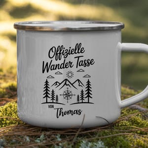 Puede incluir: Taza de camping de esmalte blanco con borde y asa plateados. La taza presenta un gr&aacute;fico negro con el texto "Offizielle Wander Tasse" y un dise&ntilde;o de br&uacute;jula, monta&ntilde;as, &aacute;rboles y el nombre "Thomas".