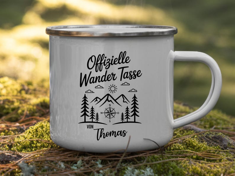 Puede incluir: Taza de camping de esmalte blanco con borde y asa plateados. La taza presenta un gr&aacute;fico negro con el texto "Offizielle Wander Tasse" y un dise&ntilde;o de br&uacute;jula, monta&ntilde;as, &aacute;rboles y el nombre "Thomas".
