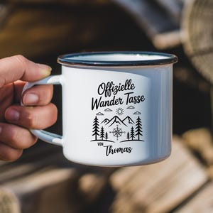 Puede incluir: Taza de camping de esmalte blanco con borde y asa negros. La taza presenta el texto "Offizielle Wander Tasse" con un dise&ntilde;o de monta&ntilde;a, br&uacute;jula y &aacute;rboles. El nombre "Thomas" est&aacute; impreso debajo.