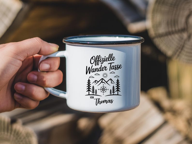 Puede incluir: Taza de camping de esmalte blanco con borde y asa negros. La taza presenta el texto "Offizielle Wander Tasse" con un dise&ntilde;o de monta&ntilde;a, br&uacute;jula y &aacute;rboles. El nombre "Thomas" est&aacute; impreso debajo.